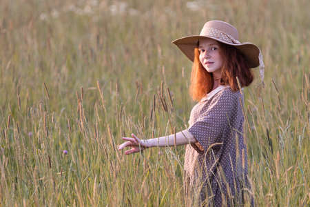 emotional redhead girl playing in the grass on a summer meadow at sunset in an elegant dressの写真素材