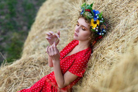 Female outdoors. A young pretty woman in a red dress posing on haystack background. Concept photo shot in a wheat field late summer eveningの写真素材