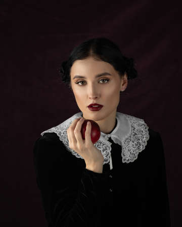 Studio portrait of pretty woman dark hair, white collar. Face, Thinking. Hold red apple. Dark backgroundの写真素材