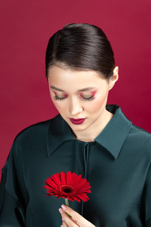 Lovely girl wearing green dress standing isolated over red background, posing with a herbera flowerの写真素材