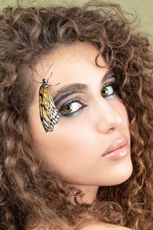 Beautiful young woman with perfect skin and bright yellow make up posing with butterfly on her face. beauty model with color professional makeup. SPA, wellness, make-up, skincare concept.の写真素材