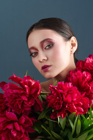 Beautiful girl with flowers peonies in hands on a blue background. Joyful female model posing in studio with bouquet of spring flowersの写真素材