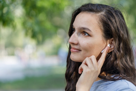 young brunette female touching TWS earbuds while listening to music during outdoor walkの写真素材