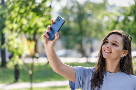 caucasian girl teenager student useaing smartphone taking selfie outdoors in city parkの写真素材