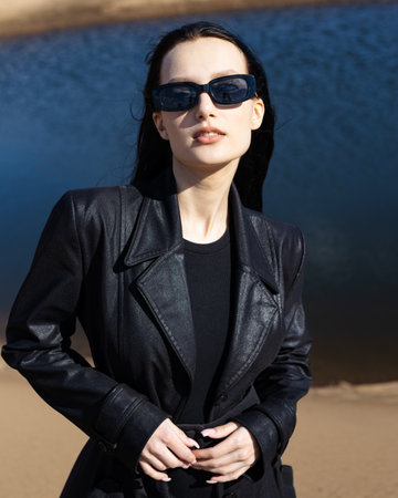 Caucasian young pretty stylish woman near river lake water street on sand pier dressed brown trench coat hold handbag smiling poses outside city, spring autumnの写真素材