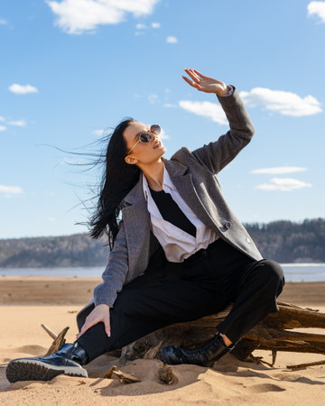 young brunette woman sitting on a sandy beach and getting rest. beautiful woman wearing fashion wardrobe looking aside to the river. warm autumn weekendの写真素材