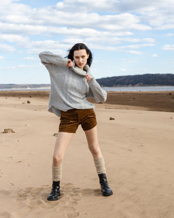 young brunette woman sitting on a sandy beach and getting rest. beautiful woman wearing fashion wardrobe looking aside to the river. warm autumn weekendの写真素材