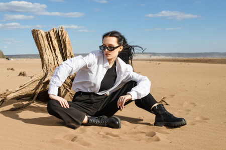 young brunette woman sitting on a sandy beach and getting rest. beautiful woman wearing fashion wardrobe looking aside to the river. warm autumn weekendの写真素材