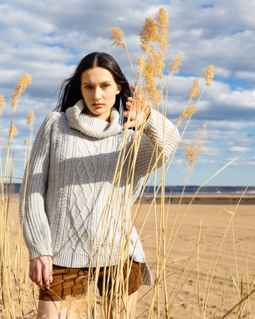 young brunette woman sitting on a sandy beach and getting rest. beautiful woman wearing fashion wardrobe looking aside to the river. warm autumn weekendの写真素材