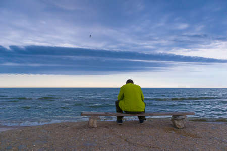 Man sitting on a bench near a beach and looking on the ocean with epic clouds.の写真素材
