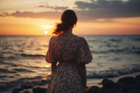 A young woman in a long dress stands on the seashore at sunset.の素材