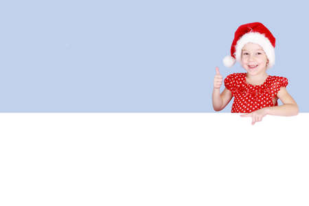 In a red dress and hat of Santa Claus a little girl shows a finger on a billboardの写真素材