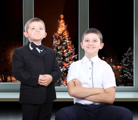 The younger brother in a suit and with a butterfly on her neck, and older brother in a shirt on a background of christmas trees outside the windowの写真素材