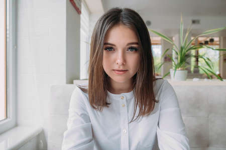 Young girl of Caucasian appearance in a white shirt sits in a cafe by the window, looking into the camera.の写真素材
