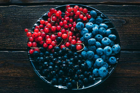 Red black currants and blueberries in a black ceramic plate on a dark wooden background in the...
