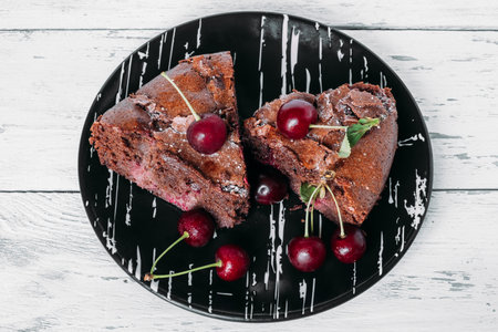 Two pieces of brownie brownie on a black plate sprinkled with cherries. Plate with cake on a white...