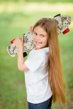 Portrait of a girl with long hair holding a painted skateboard on her shoulder in the park. Girl smiling at the cameraの写真素材