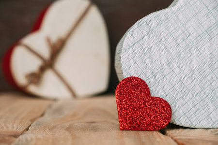 Valentine's Day. Red shiny small heart on the background of gift boxes in the form of hearts on a wooden table.の写真素材