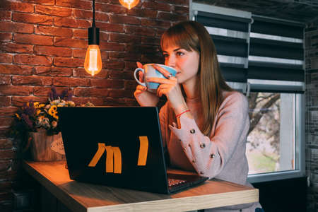 Remote work at home. The girl sits at home in the kitchen and uses a laptop. He holds a cup of tea in his hands and looks forward.の写真素材