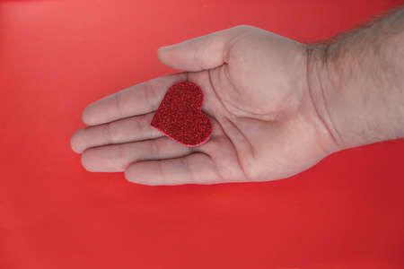 Small red shiny heart on man's palm on red background. Declaration of love, expression of feelings. Copy, empty spaceの写真素材