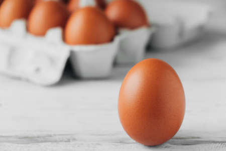 Brown chicken egg standing on wooden table. In background is cardboard tray filled with eggs....