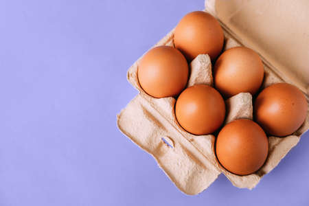 Top view of an open cardboard box with brown eggs on a purple background. A popular all-purpose food...