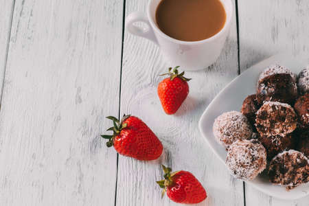Brownie cakes in coconut flakes on plate. Nearby is white cup with coffee and fresh strawberries. copyspace.の写真素材