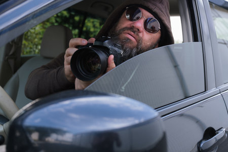 A man with a beard, wearing a hood and sunglasses, takes photographs from a car window. His focused gaze and reflection in the glass create an atmosphere of observation and mysteryの写真素材