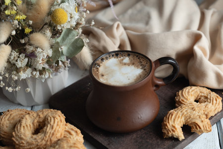 A cup of cappuccino in a ceramic brown mug on a wooden board. Nearby lie crumbly cookies in the shape of curls. The background is beige textiles and a vase of dried flowersの写真素材