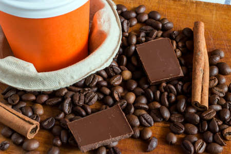 Coffee in orange paper glass with background of coffee beans, chocolate chunks and cinnamon sticks