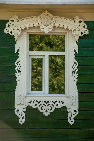 A window with a wooden frame and carved ornaments on the background of a green wall of boards.の写真素材