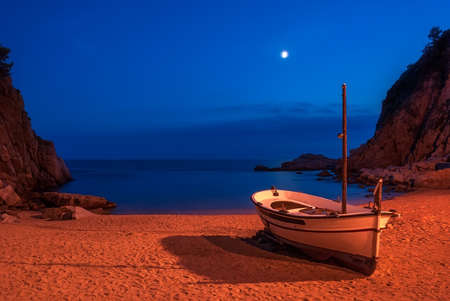 A boat on a sandy beach in Tossa De Mar Bay lunar night against the backdrop of mountains and blue skies.の写真素材
