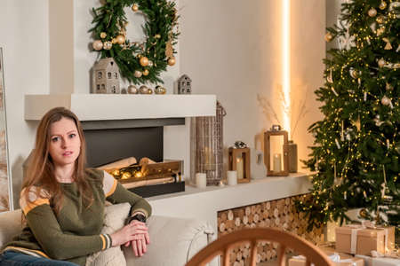 A young, beautiful woman is sitting on a sofa in a living room with a Christmas interior. New Year's mood and interior with a Christmas tree decorated with a garland.の写真素材