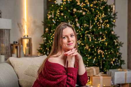 A young, beautiful woman is sitting on a sofa in a living room with a Christmas interior. New Year's mood and interior with a Christmas tree decorated with a garland.の写真素材