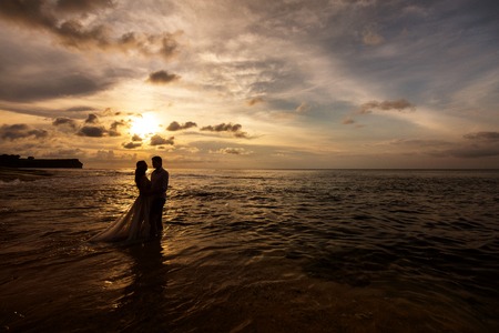 Silhouettes of bride and groom embracing standing in the sea water on the beach at sunsetの写真素材