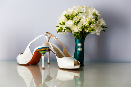 Bridal bouquet of white freesias, decorated with satin ribbon on the background of the brides high heel shoesの写真素材