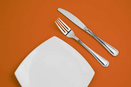 Knife, square white plate and fork on orange backgroundの写真素材