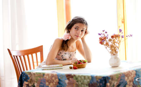 Pensive girl with book sitting at table indoor in summer day with strawberriesの写真素材