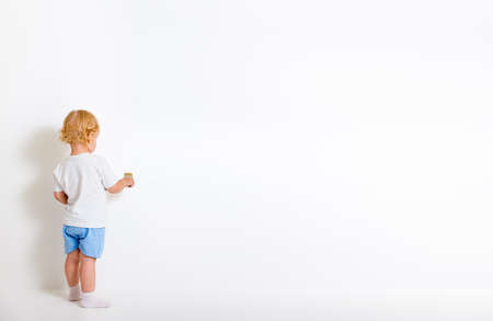 Little boy with paint brush standing back near white wallの写真素材