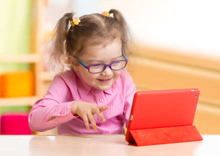 Smart kid in spectacles using tablet PC or e-book sitting at table in her roomの写真素材