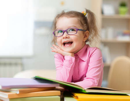 Smart kid girl in glasses reading books sitting at table at her roomの写真素材
