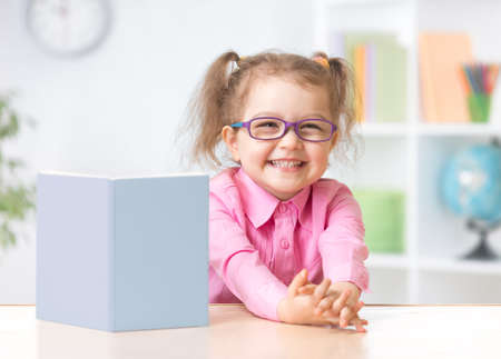 Happy kid girl in eyeglasses reading with standing book on her tableの写真素材