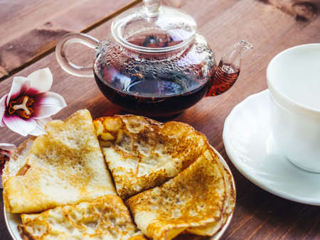 A plate with pancakes and a white cup and a teapot with tea on a wooden tableの写真素材