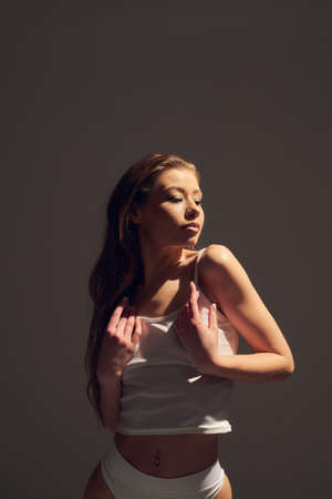 Portrait of young attractive woman model with brown long curly hair, in white bodysuit dress , long legs with tan skin, slim body, sitting on background with sun hard light. The concept of a beautiful elegant figure, body positive, healthy lifestyleの写真素材