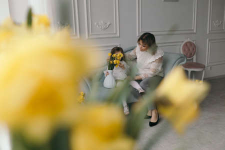 Mom and daughter in linen dresses are spinning, laughing and having fun on a large bed near a bouquet of narcissus flowerの写真素材