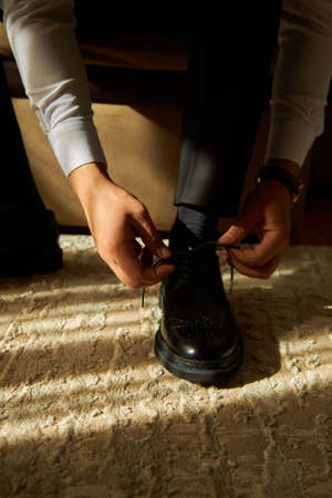 The groom ties the laces on his shoes in the morning in a sunny roomの写真素材