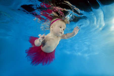 A little girl in a red skirt and crown swims underwater in the poolの写真素材