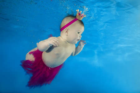 A little girl in a red skirt and crown swims underwater in the poolの写真素材