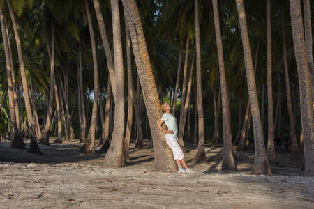 Young beautiful woman standing in a tropical forest leaning on a palm tree