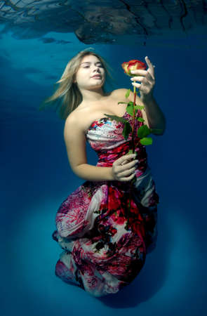 Young beautiful woman in a bright dress with a rose in hands underwaterの写真素材